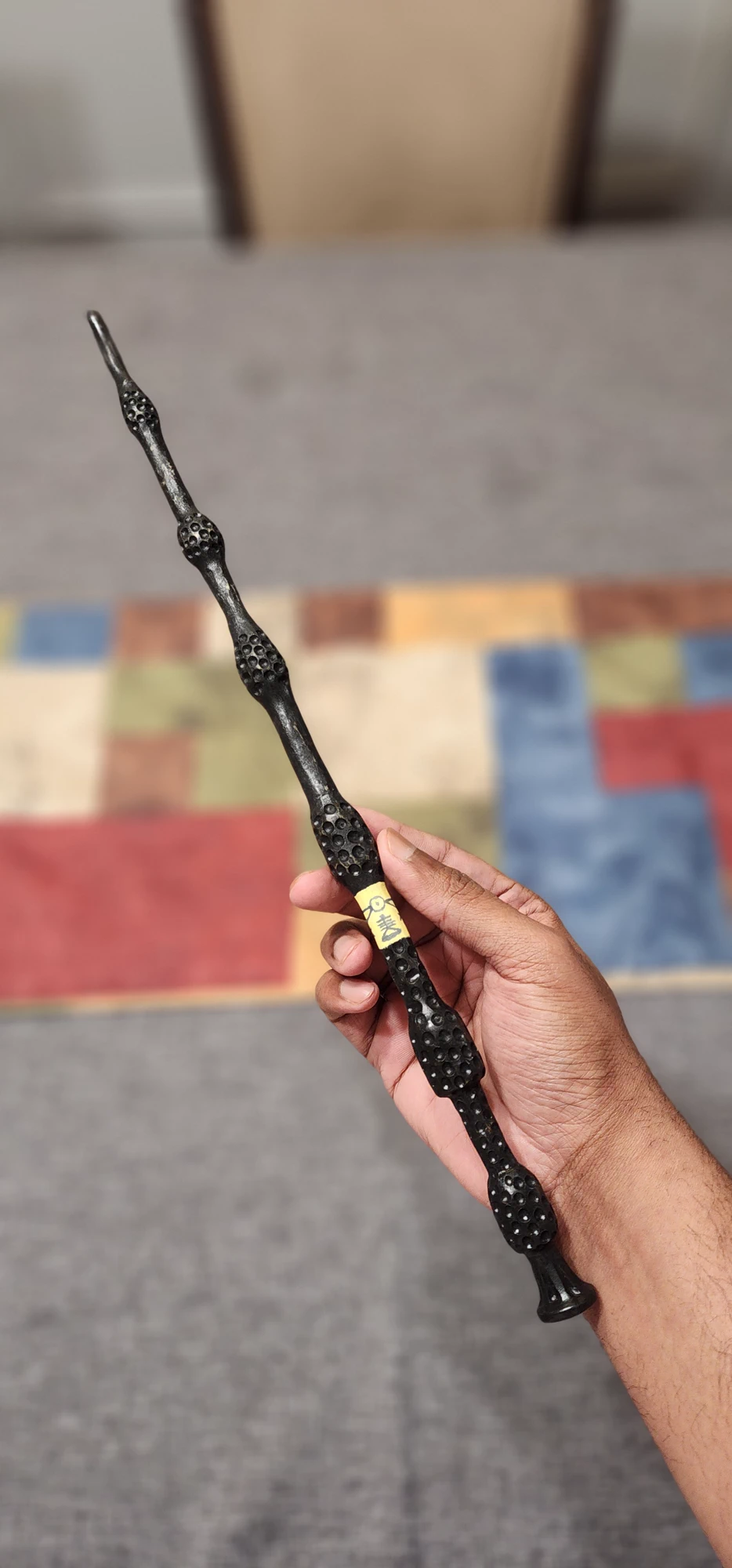 A hand holding a carved black Elder Wand above a brightly tiled pavement.